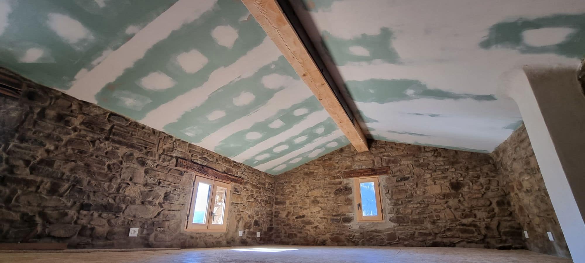 Pièce intérieure aux murs de pierre dégagés, linteaux de bois anciens au-dessus des fenêtres, plafond neuf et poutre apparente.
