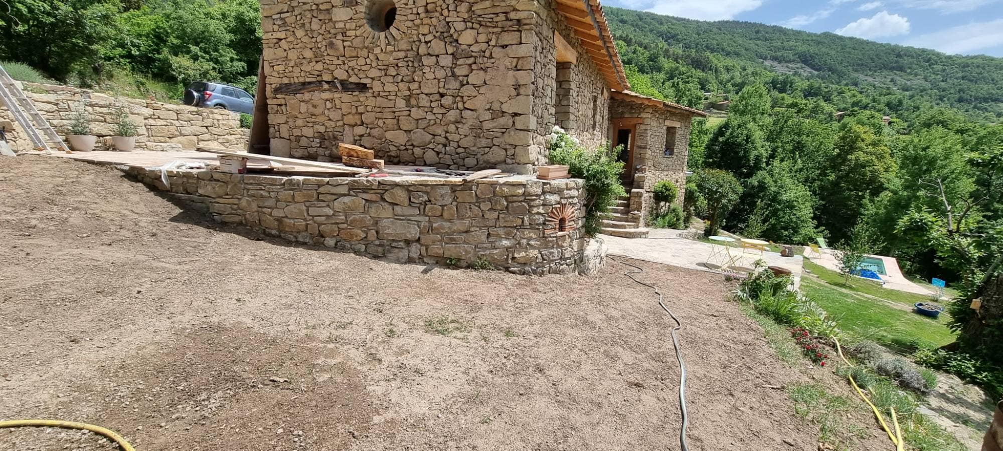 Mur de soutènement courbe en pierre sèche portant la terrasse de la bâtisse, face à la vallée boisée.