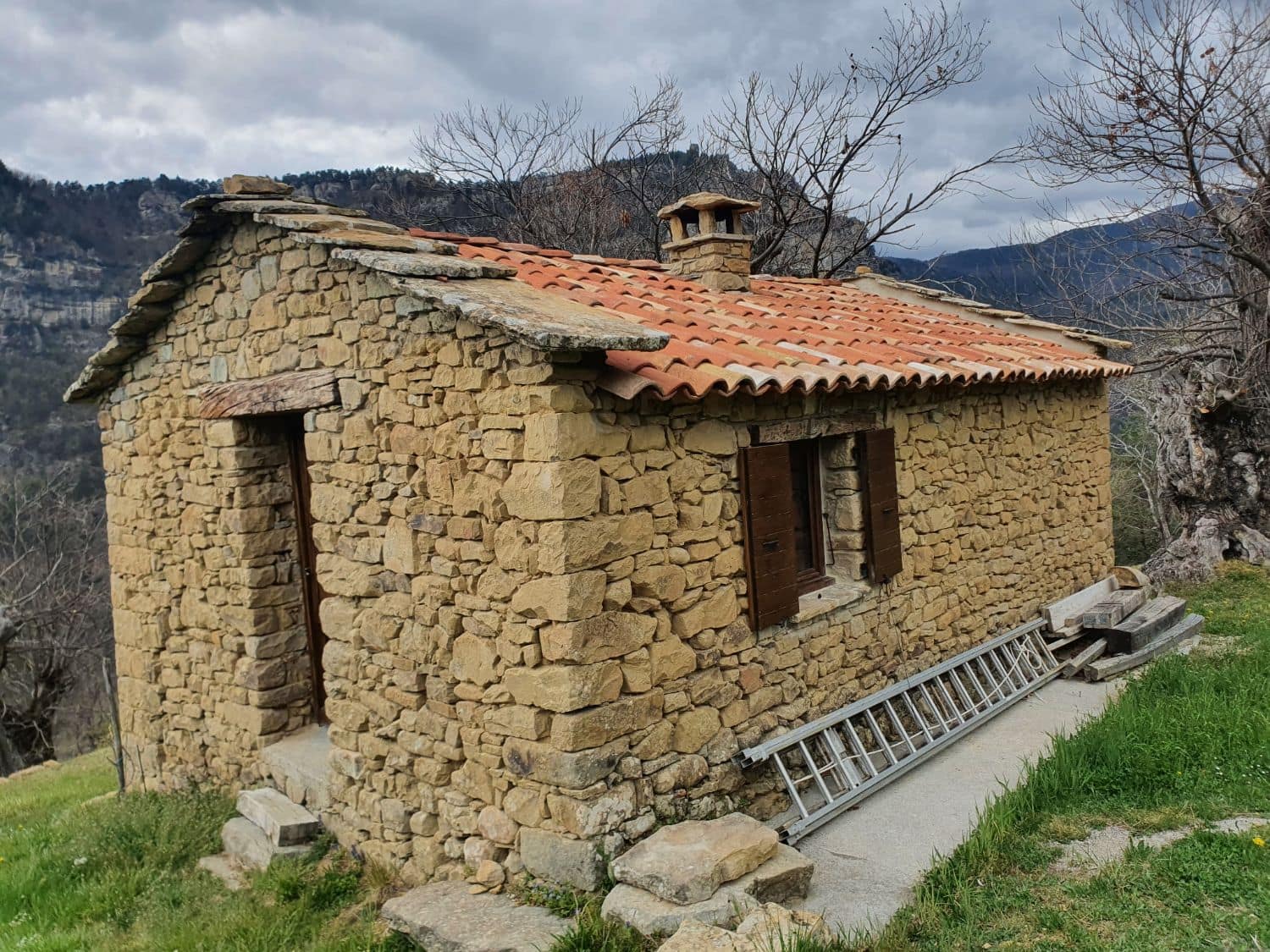 Cabanon en pierre, couverture en tuiles et rives en lauzes, face à la montagne.