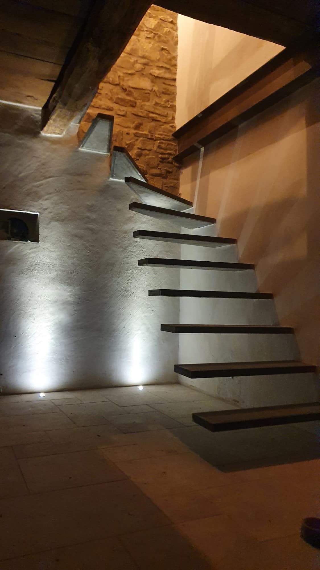 Escalier à marches suspendues contre un mur de pierre, éclairage encastré au sol.