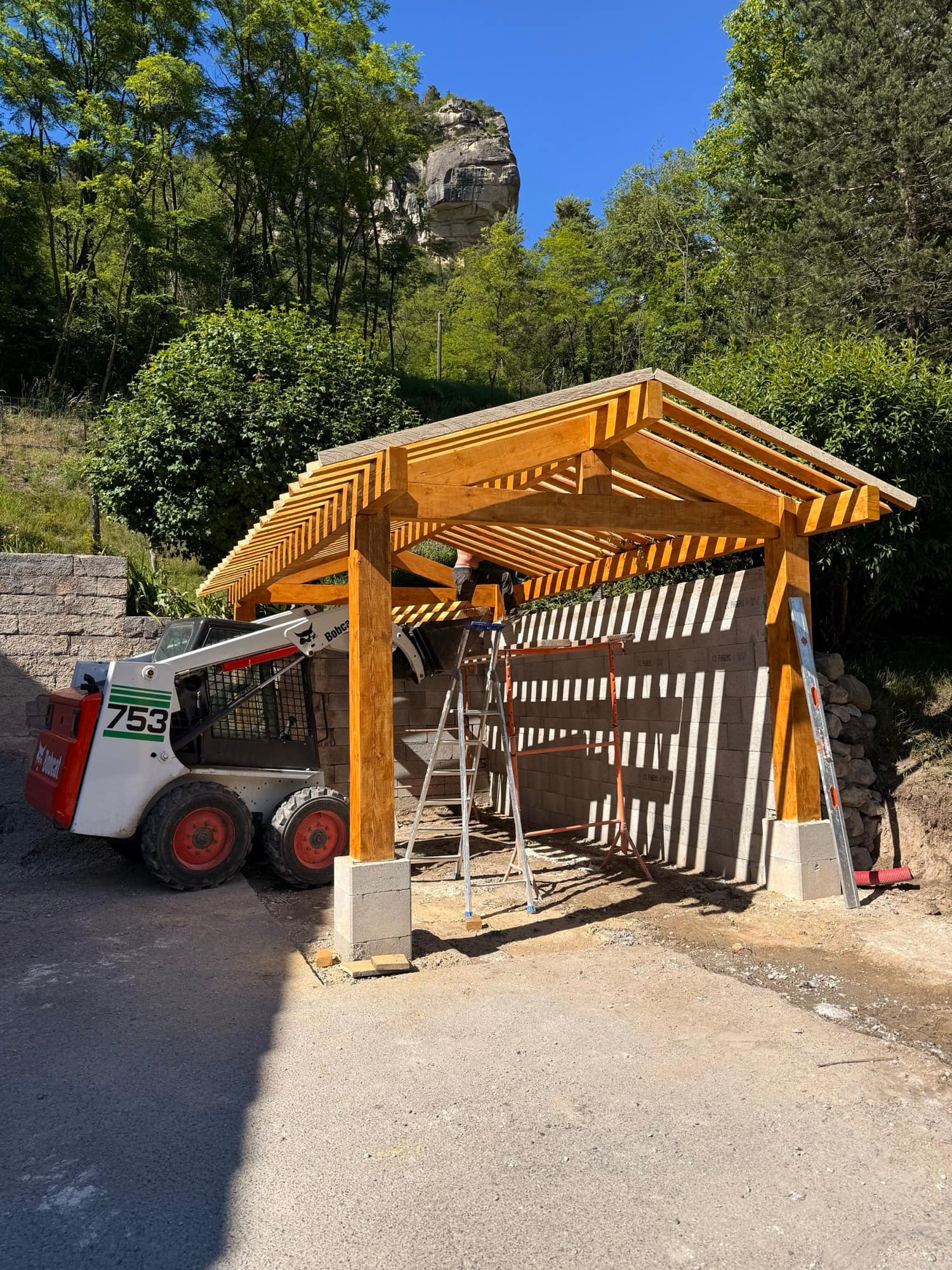 Chantier de gros œuvre : charpente en bois massif posée sur des murs en blocs, engin de chantier au premier plan.