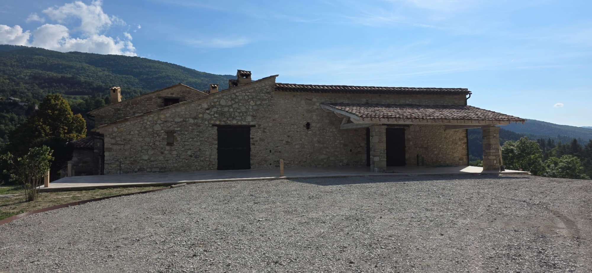 Longue bâtisse en pierre et son auvent sur piliers de pierre, toiture en tuiles refaite, cour gravillonnée face aux montagnes.