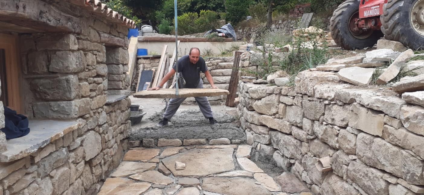 Maçon portant une dalle de pierre pour la poser, entre deux murs en pierre sèche.