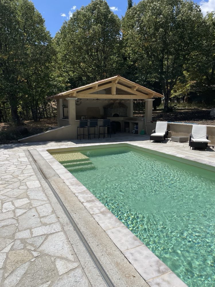 Piscine et pool house sous charpente de bois, ceinturés d’un dallage de pierre.