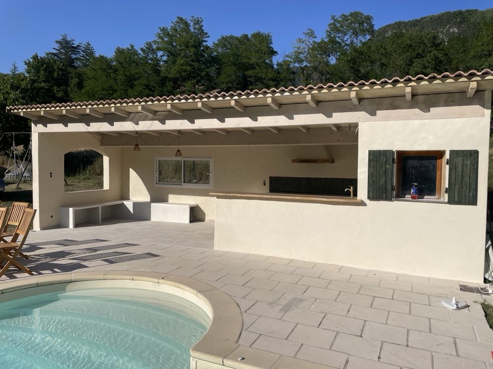 Pool house enduit et couvert de tuiles rondes, charpente de bois apparente, au bord du bassin.